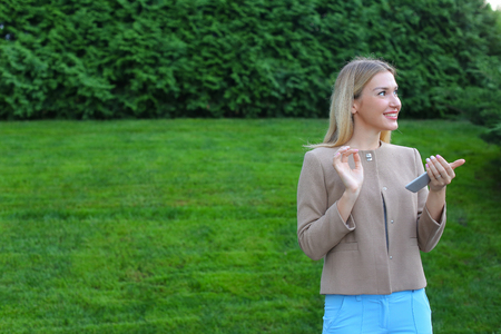 Stylish young girl of european appearance views all around and smiles little smile, thinking about something pleasant. Girl dressed in light blouse, beige jacket and blue pants, stands on background of green lawn and coniferous plants outdoors. Concept ofの写真素材