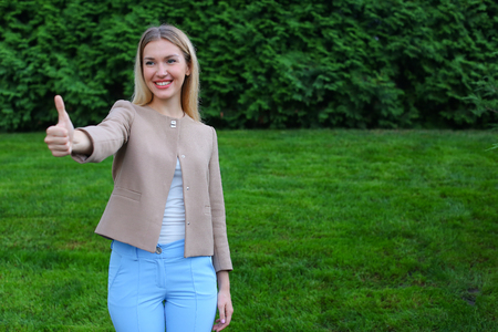 Slender blonde smiles and shows gesture of "Cool", enjoying life and new day, advertises and shows evaluation of cosmetic product, recommends beauty salon or restaurant. Women with long blonde hair  dressed in bright blouse, beige jacket and blue pants, sの写真素材