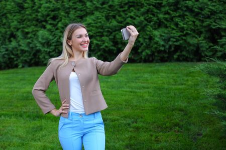 Active female blonde calls up and talking on phone, waving and smiling companion to your phone, make selfie, reported good news, and shows emotion gestures. Girl with long hairdressed in bright blouse, beige jacket and blue pants, standing on background oの写真素材