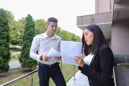 Two young educated boy and girl, successful entrepreneurs, students communicate, negotiate working moments, see into plans, kept in hands paper, graphics and view them, stand on background of office buildings and trees outdoors. Girl with dark hair dresseの写真素材