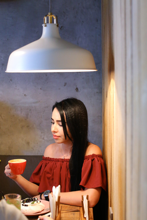 Lovely young female girl holds in hand red cup and drinks coffee behind wooden square table against background of gray wall in cozy coffee shop with gray modern lamp. On table stand desserts and holder for napkins. Girl with black hair dressed in maroon dの写真素材