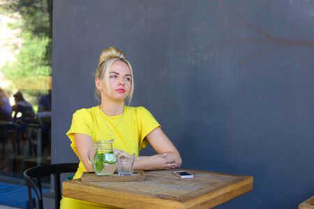 Stunning cute young girl female woman looks forward,waiting, sits at wooden square table against background of gray walls and windows in coffee shop outdoors. Girl with blond hair dressed in short citric dress.Concept of meeting with friends, female frienの写真素材