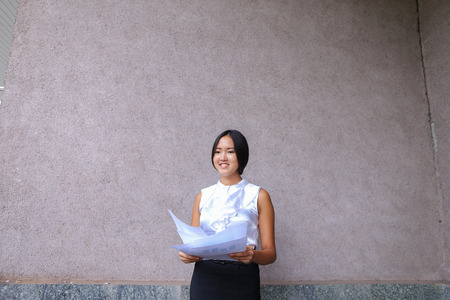 Pretty asian girl smiling and posing, holds paper in hands and looking away on background of wall of buildings outdoors. Woman with black hair, dressed in white blouse and classic skirt. Concept of business meeting, business communication, successful peopの写真素材