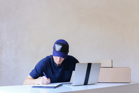 Mailman Sitting in Office on Table and Typing at Laptop, Taking Pen and Writes Information on Document. Young Student Works as Courier and Successfully Performs Work, Worker Has European Appearance in Black T-Shirt and Cap Sitting at White Table Typing onの写真素材