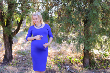 Marvelous Young Adult Female to Carry Child Under Ones Heart, Relax and Enjoy Nature and Fresh Air, and Stands in Big Green Park Outdoors in Daytime. Girl With Blond Hair, Braided in Braid in Front Dressed in Long Blue Dress. Concept of Happy Pregnancy anの写真素材