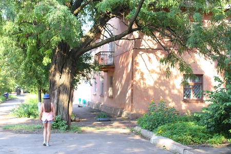 Girl child stands with his back to the photographer, goes, directed towards the house, walking. Girl dressed in pink shirt, Printed shorts, dark blue hat with a brim and blue sandals. Concept of summer holidays, childhood, walk.の写真素材