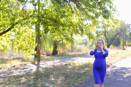 Dazzling Contemporary Young Adult Feminine of European Appearance Carries Baby Under Heart. Girl Leisurely Walking and Chatters on Phone With Husband About Well-Being and Appoint Meeting at Cinema or Says Family Doctor About State of Health. Female With Bの写真素材