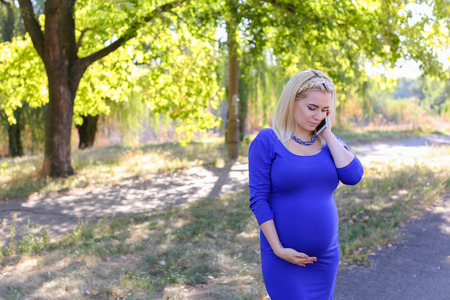 Dazzling Contemporary Young Adult Feminine of European Appearance Carries Baby Under Heart. Girl Leisurely Walking and Chatters on Phone With Husband About Well-Being and Appoint Meeting at Cinema or Says Family Doctor About State of Health. Female With Bの写真素材