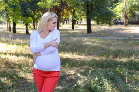 Lovely Woman and Future Mother Prepares For Birth of Baby, Tired and Trying to Relax, Posing For Photos on Memory and Stands in Green Sunny Park Outdoors in Daytime. Girl With Blond Hair, Braided in Braid in Front Dressed in White Blouse With Long Sleevesの写真素材