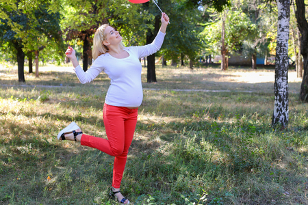 Alluring Future Mother of European Appearance Enjoys Pleasant Things and Look Forward to Appearance of First-Born Into World Photographed For Advertising of Womens Clothing For Pregnant With Pink Ballon in Shape of Heart in Green Sunny Park Outdoors in Daの写真素材