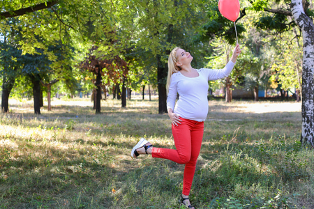 Alluring Future Mother of European Appearance Enjoys Pleasant Things and Look Forward to Appearance of First-Born Into World Photographed For Advertising of Womens Clothing For Pregnant With Pink Ballon in Shape of Heart in Green Sunny Park Outdoors in Daの写真素材
