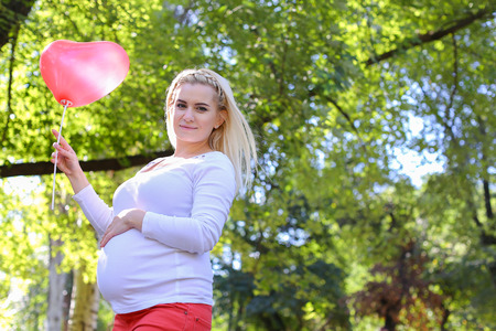 Marvelous Young Adult Lady With Child of European Appearance Enjoying Sunny Day, Cute Smiles, Poses and Rises up Pink Ballons in Shape of Heart in Green Park Outdoors in Daytime. Female With Blonde Hair Braided in Braid in Front Dressed in White Blouse Wiの写真素材