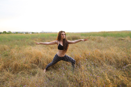 Young female fitness instructor performs warm-up exercise, stretching and holding focuses on correct execution poses, photographed for advertising of catalog womens sportswear or sports complex in wide field outdoors. Sportswoman with curly brown hair dreの写真素材