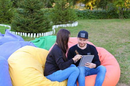 Pair of lovers of young attractive people sitting in soft yellow and red chair, man uses gadget that keeps in arms, female holds plastic card, coupon. Young people cute communicate, plan to travel, choosing interior of apartment, she shows on flyer and guの写真素材