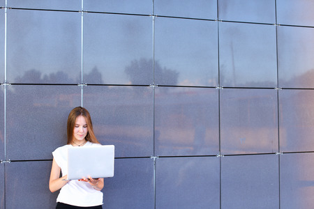 Beautiful young female woman girl entrepreneur successful person holds laptop in hands works, solves problems, and stands on background wall of modern business center.Girl with long hait dressed in white shirt and black trousers.Concept of business meetinの写真素材