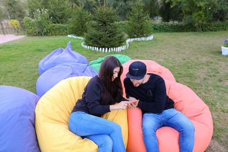 Modern beautiful enamored people, beautiful girl and young man sitting in soft yellow and red chair, planning journey, girl holds phone and talking with boyfriend, booked hotel for total travel, young people smile at each other, located in large green parの写真素材