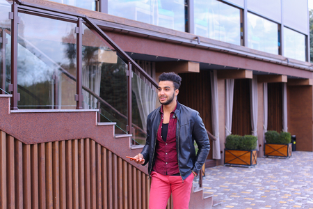 Handsome Young Adult Muslim Man, Businessman Walks Slowly Along Stairs, Looks Around and Smiles, Holds Hands in Pockets of Jackets, Advertises Clothing on Background of Wooden Stairs Near Restaurant Outdoors. Confident Arab Guy With Dark Hair Dressed in Bの写真素材