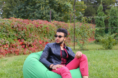 Arabian Young Man Sitting and Resting From City Bustle in Soft Green Chair, smiles and Looks Around. Handsome Guy Has Dark Hair and Black Beard and dressed in Maroon Shirt Over Black Leather Jacket and Bright Pink Jeans and White Sneakers Accessories Fromの写真素材