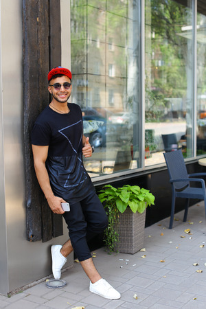 Handsome young man arab stands near wall smiles, rejoices and holds in hand mobile phone on background of panoramic windows in modern restaurant. Man dressed in dark T-shirt, black pants, white sneakers and black red color cap and sunglasses. Concept of lの写真素材