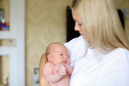 Young mother gently hugging, swaying and talking, singing song to newborn, child looks at mother and looks around, located mothers arms in bright room. Woman blonde European appearance dressed in white T-shirt. Concept of maternal love and care for baby, の写真素材