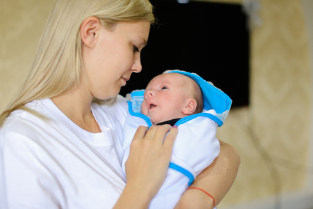 Young female lulls and sings songs for child, gently hugging and talking with kid, baby cries and looks at mother, located in mothers arms in bright room. Woman blonde European appearance dressed in white T-shirt, newborn baby wrapped in white blanket witの写真素材