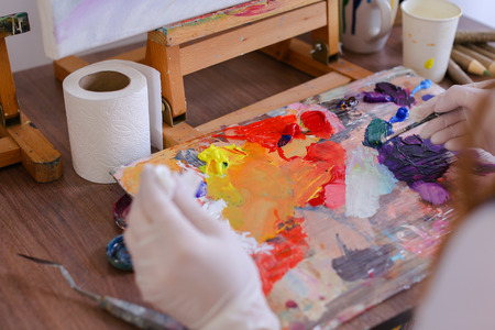 Women Artist extrudes hands in white gloves oil paint from tubes on multi-colored palette for viewing and mixing colors to start drawing pictures in art studio. On table stands glass of water, jar of colored pencils and brushes and palette sketch. Conceptの写真素材