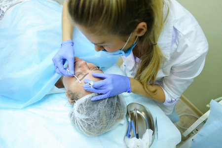 Master of mikrobleyding young attractive girl evaluates correct execution of work, check with help of special device density of drawing and color of hairs of eyebrows, brings to end of last stage in bio tattooing eyebrows. Master dressed in white robe, blの写真素材