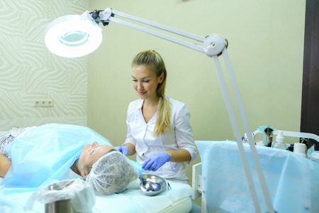 Blonde girl European appearance, professional engaged in reconstruction of eyebrows responsibly preparing to start work mikrobleyding eyebrows, eyebrows customer focused prepares women to paint application, choose appropriate shade for tattoo, next to couの写真素材