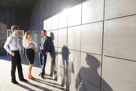 Contemporary perspective elegant young people, two boys and a girl, businessmen and students to meet and discuss important issues, share business ideas, smiling, laughing, make decisions, solve problems and standing near gray wall of modern business centeの写真素材