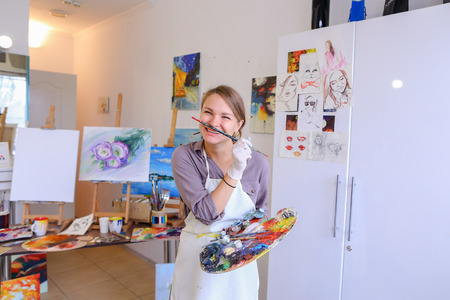 Cute female painter posing, laughing and fooling around in front of camera and smiling with brushes and colorful palette in hand, standing in studio with white art paintings on walls. Girl European appearance advertises art school for adults and children,の写真素材