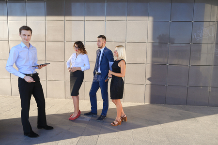Handsome confident young male guy, businessman, student holds tablet in hands and deliver presentations, talks about gadget, advertises new technology to colleagues who listening to attentively, and stand on background wall of modern business center outdoの写真素材