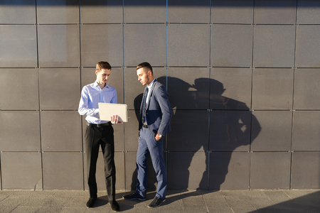 Handsome confident young male man, businessman, student holds laptop in hands and deliver presentations, talks about gadget, advertises new technology to colleague who listening to attentively, and stand on background wall of modern business center outdooの写真素材