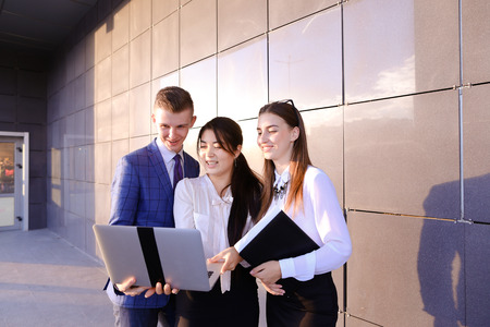 Modern young man and two attractive smart young women, business people, students communicate, holding laptop and study work plan, familiarize with presentation, teach each other new programs, solve important problems and workloads holding folder with docuの写真素材