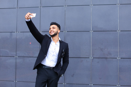 Entrepreneur takes makes selfie man male looking at camera and smiling talks on mobile smart phone. Young handsome businessman arab muslims in business center wearing dressed in black elegant suit on building background.の写真素材