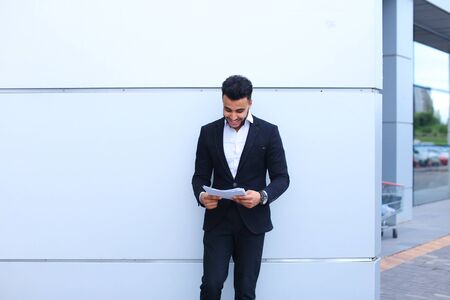 Young adult successful student businessman arab man holds documents, securities, smiling and thinks near gray wall of business center. Muscular bearded man dressed in classic suit, black pants, jacket , white shirt, on hand is watch. Concept of securitiesの写真素材