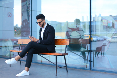 Entrepreneur smiling man  puts on sunglasses uses tablet and looks at. Young handsome businessman arab muslims in business center wearing dressed in black elegant suit on building background.の写真素材