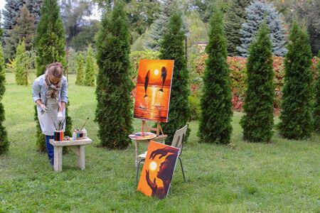 Artist cleans workplace, evaluate work that stands on easel, smiling, posing and looking in camera, standing in full length near paintings in park outdoors on background of coniferous plants in afternoon. Girl dressed in red sweater, dark jeans, white aprの写真素材