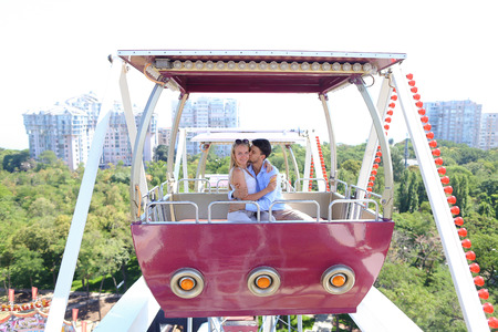 Enamored couple of beautiful people, boy and girl kissing, hugging, posing for photo, ride on ferris wheel, smiling, laughing and sit in booth ferris wheel on background of city, sky and trees at an amusement park outdoors. Girl with long blond hair dressの写真素材