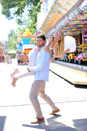 Sweethearts cuddling, pose for camera and fooled, boy holding girl with hand on shoulder at an amusement park outdoors. Girl with long blond hair dressed in white top and beige pants, with small blue bag on shoulder, guy dressed in blue light shirt and beの写真素材