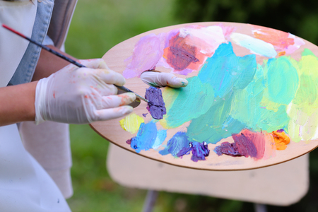 Photo of female hand artist who mixes oil or acrylic colored bright colors on wooden panel, which would start to draw and located in green park in open air. Concept of set of objects to paint, oil or acrylic paints, advertising art school.の写真素材