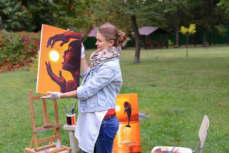 Female European appearance artist evaluates picture and removes from easel, holds and posing with picture which depicts woman with sun in hands made orange smiling and standing in full growth on background of lawn and angered by conifers in large park in の写真素材