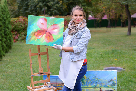 Female European appearance artist evaluates picture and removes from easel, holds and posing with picture which depicts colorful butterfly, smiling and standing in full growth on background of lawn and angered by conifers in large park in open air. Girl dの写真素材