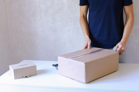 Deliveryman Standing in Room, Glue Tape Box and Takes Parcel in Hands and Smiling . Postman European Appearance in a Black T-Shirt and Cap Stands Near the Desk in the Office With White Walls and Unassisted Packaging Boxes, Adhesive Glue Tape on a Cardboarの写真素材