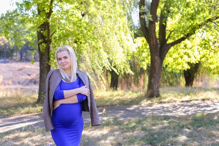 Charming Lady Being Prepared to Become Mother. Young Woman Photographed For Album of Family Photographs or Advertising Clothes For Pregnant Women, Waiting For Her Husband in Park to go Along to the Restaurant For Dinner. Blonde With Hair Braided in Pigtaiの写真素材