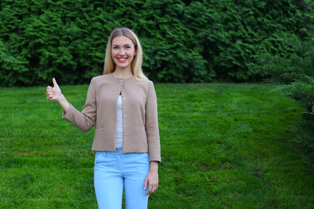 Slender blonde smiles and shows gesture of "Cool", enjoying life and new day, advertises and shows evaluation of cosmetic product, recommends beauty salon or restaurant. Women with long blonde hair  dressed in bright blouse, beige jacket and blue pants, sの写真素材