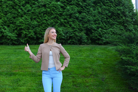 Slender blonde smiles and shows gesture of "Cool", enjoying life and new day, advertises and shows evaluation of cosmetic product, recommends beauty salon or restaurant. Women with long blonde hair  dressed in bright blouse, beige jacket and blue pants, sの写真素材