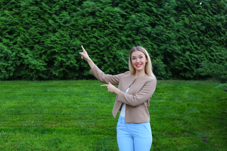 Girl gestures and shows fingers on certain place in picture, advertise any product, smiles and stands on background of green plants outdoors. Women with long blonde hair dressed in bright blouse, beige jacket and blue pants. Concept of emotional expressioの写真素材