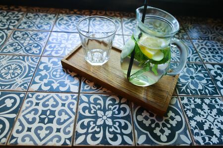 Glass decanter and black drinking straw with refreshing drink from cucumber, lemon, mint and empty glass faceted cup stands on tile with patterns in cafe. Concept of refreshing drink, non-alcoholic cocktails, restaurants business, menu of beverages.の写真素材