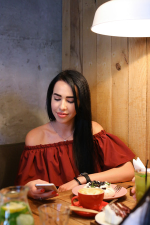 Cute female girl smiles, holds in hands phone and uses it, conducts correspondence, solves questions dials sms at table in cafe. On table stand plates of confectionery, meringue in pink round plate and cherry pie, red cup with coffee drink, latte, cappuccの写真素材