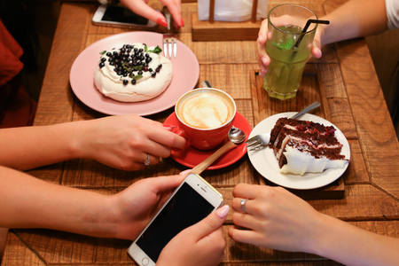 Girls met in cafe and ordered delicious pastry, cherry pie on white circular plate, meringue with black currants on round pink plate, refreshing lemonade in glass jar of cucumber, lemon and mint and cup of coffee beverage, latte with pattern on cream in rの写真素材